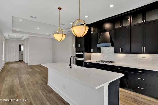 kitchen featuring dark cabinets, custom exhaust hood, stainless steel appliances, light wood-type flooring, and a sink