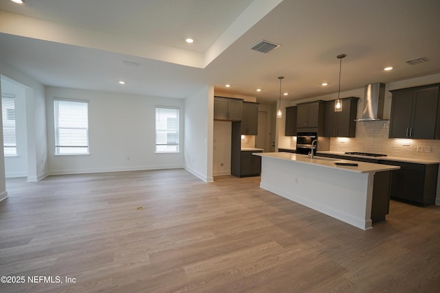 kitchen featuring wall chimney exhaust hood, an island with sink, pendant lighting, light hardwood / wood-style floors, and black appliances