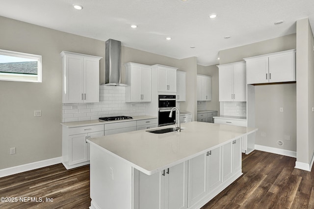 kitchen featuring a kitchen island with sink, a sink, wall chimney range hood, dark wood-style floors, and multiple ovens