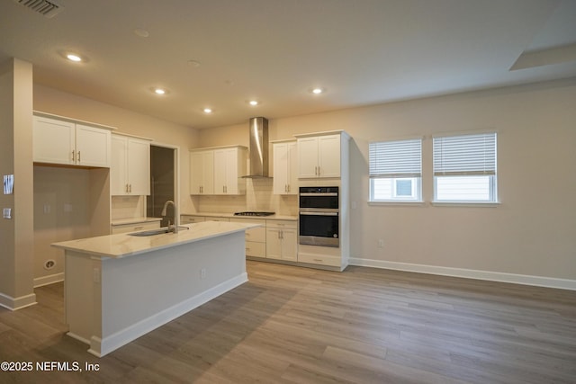 kitchen with stainless steel double oven, a kitchen island with sink, sink, wall chimney range hood, and white cabinetry