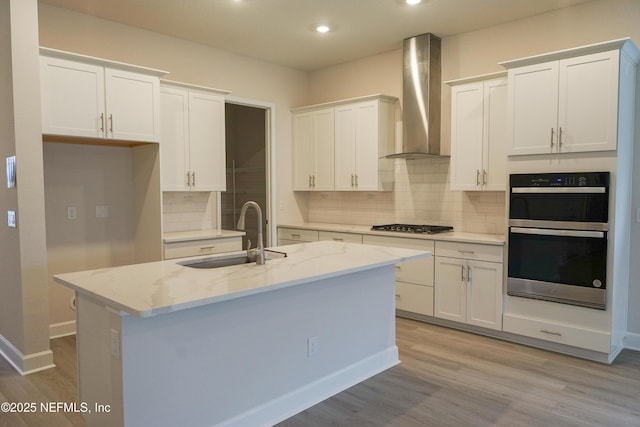 kitchen with appliances with stainless steel finishes, wall chimney exhaust hood, sink, a center island with sink, and white cabinets