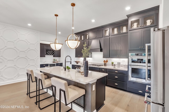 kitchen featuring light stone counters, a kitchen island with sink, stainless steel appliances, wall chimney range hood, and light wood-type flooring