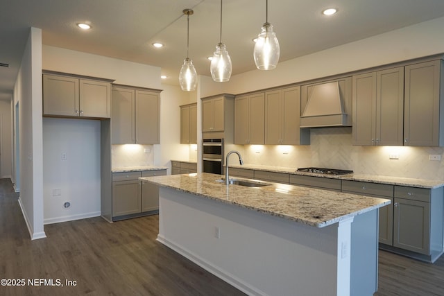 kitchen featuring light stone countertops, sink, premium range hood, a center island with sink, and appliances with stainless steel finishes