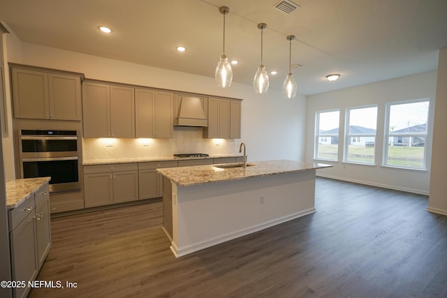 kitchen featuring sink, stainless steel appliances, light stone counters, an island with sink, and custom exhaust hood