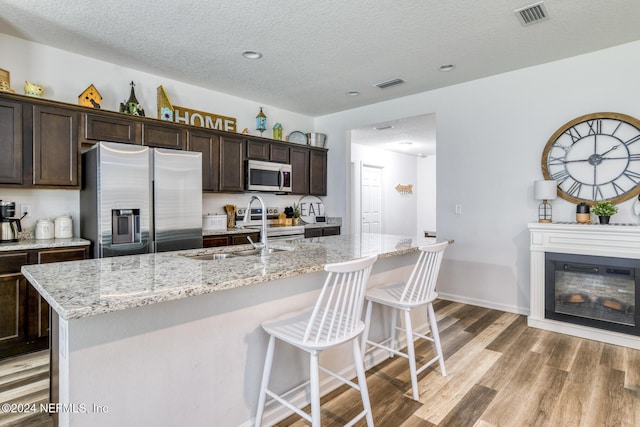 kitchen featuring light wood-type flooring, a textured ceiling, dark brown cabinetry, stainless steel appliances, and a kitchen island with sink