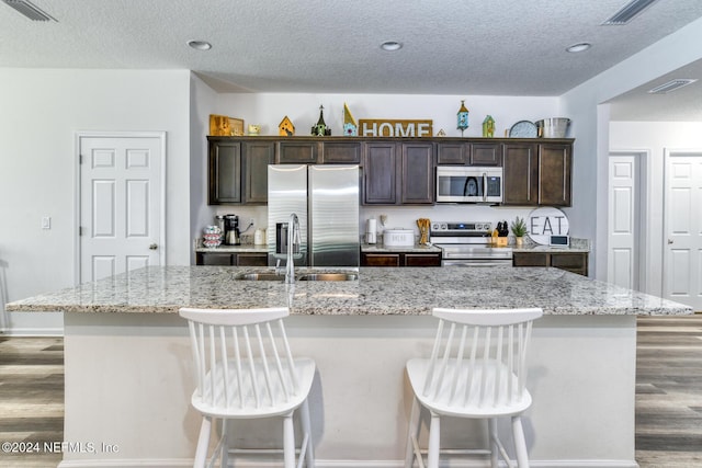 kitchen with dark brown cabinetry, light stone countertops, stainless steel appliances, a kitchen island with sink, and a breakfast bar