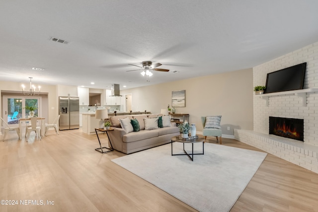 living room featuring a textured ceiling, ceiling fan with notable chandelier, light hardwood / wood-style flooring, and a brick fireplace