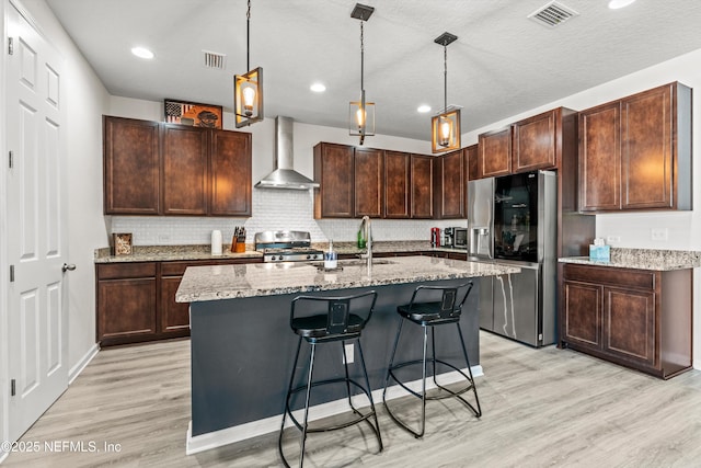 kitchen with pendant lighting, a kitchen island with sink, wall chimney exhaust hood, light stone countertops, and stainless steel appliances