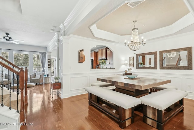 dining space with ceiling fan with notable chandelier, wood-type flooring, ornate columns, a raised ceiling, and crown molding