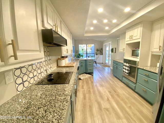 kitchen with stainless steel oven, a raised ceiling, black electric cooktop, white cabinets, and exhaust hood