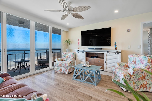 living room with ceiling fan, a water view, light hardwood / wood-style floors, and a wall of windows