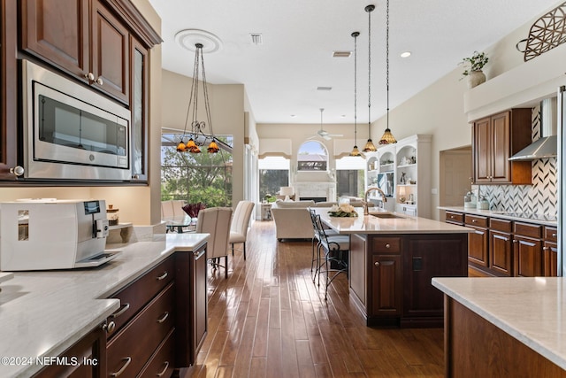 kitchen featuring ceiling fan with notable chandelier, stainless steel microwave, sink, hanging light fixtures, and an island with sink