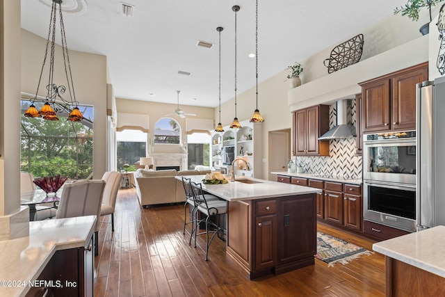 kitchen with backsplash, a center island with sink, hanging light fixtures, wall chimney exhaust hood, and stainless steel appliances