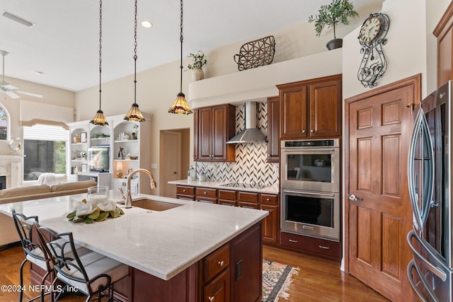 kitchen with a kitchen island with sink, wall chimney exhaust hood, ceiling fan, decorative backsplash, and appliances with stainless steel finishes