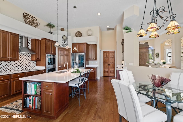 kitchen with wall chimney range hood, sink, an island with sink, appliances with stainless steel finishes, and tasteful backsplash