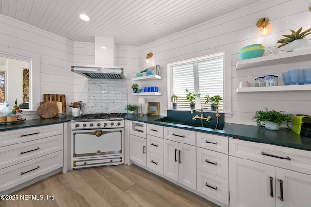 kitchen with wooden walls, wall chimney range hood, white range with gas cooktop, light hardwood / wood-style floors, and white cabinetry