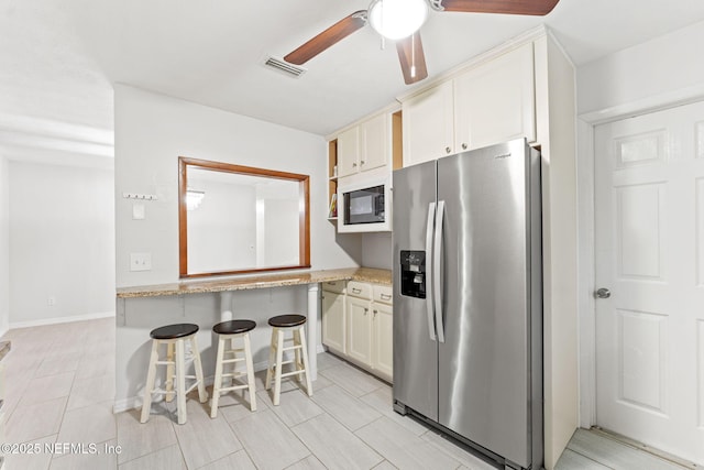 kitchen with black microwave, ceiling fan, light stone countertops, stainless steel refrigerator with ice dispenser, and a breakfast bar