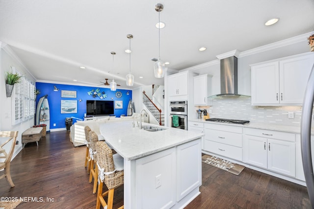 kitchen featuring a kitchen island with sink, hanging light fixtures, wall chimney exhaust hood, ceiling fan, and white cabinetry