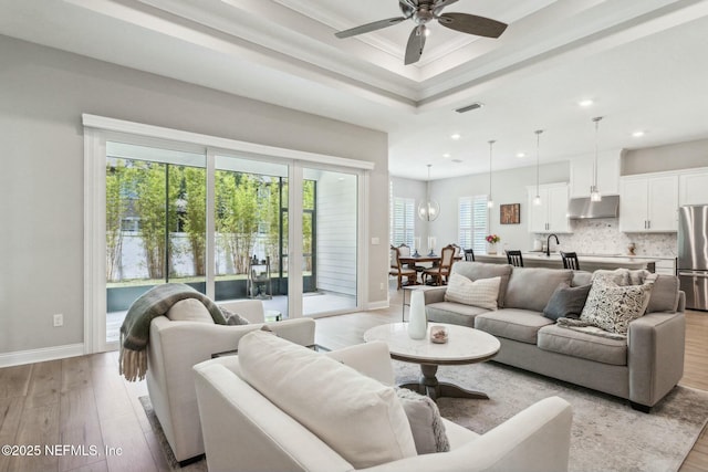 living room with ceiling fan, sink, light hardwood / wood-style flooring, a tray ceiling, and ornamental molding