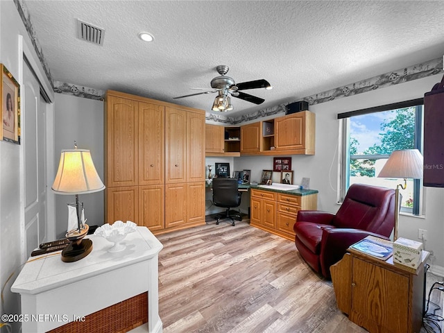 home office with light wood-type flooring, a textured ceiling, ceiling fan, and built in desk