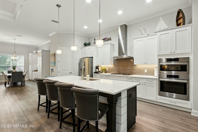 kitchen with appliances with stainless steel finishes, wall chimney exhaust hood, decorative light fixtures, white cabinets, and an island with sink