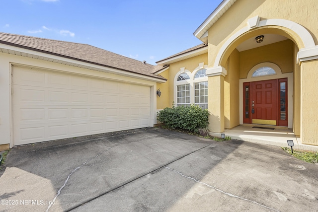 doorway to property featuring a shingled roof, driveway, an attached garage, and stucco siding