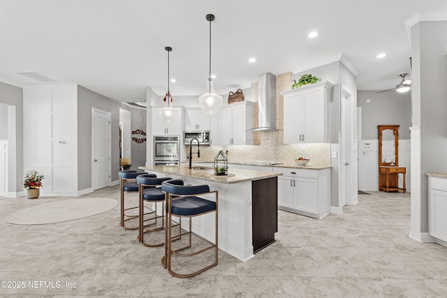 kitchen featuring hanging light fixtures, wall chimney range hood, light stone counters, an island with sink, and white cabinets