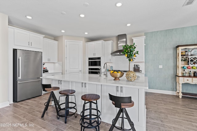 kitchen featuring wall chimney range hood, appliances with stainless steel finishes, an island with sink, white cabinets, and a kitchen bar