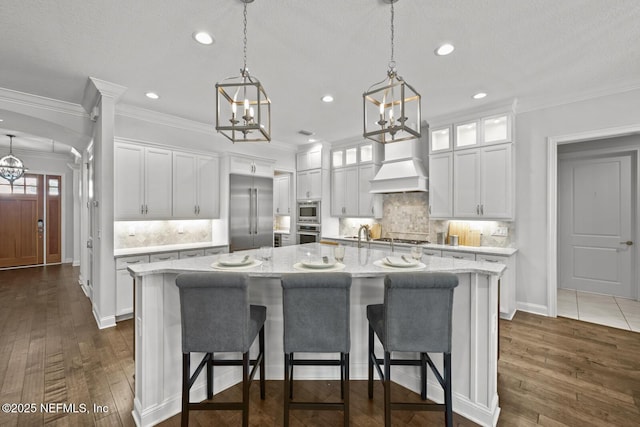 kitchen featuring white cabinetry, premium range hood, built in appliances, a spacious island, and decorative light fixtures