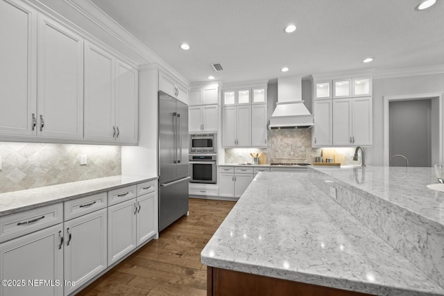 kitchen with built in appliances, light stone counters, white cabinets, and custom range hood