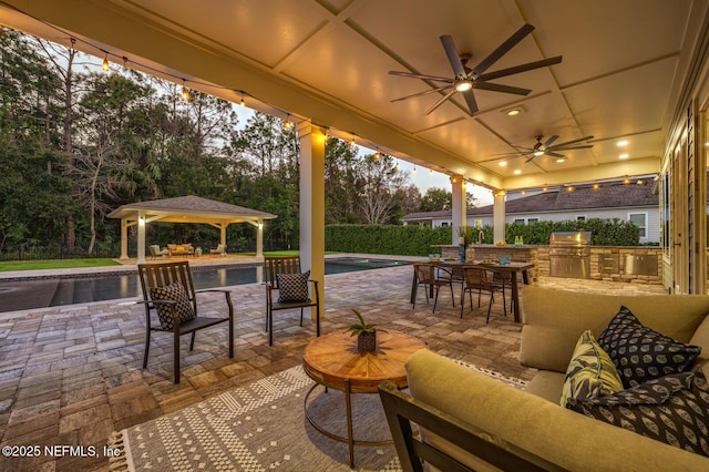 patio terrace at dusk with grilling area, an outdoor hangout area, a gazebo, ceiling fan, and exterior kitchen