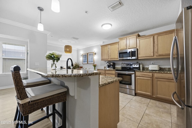 kitchen with dark stone countertops, crown molding, hanging light fixtures, appliances with stainless steel finishes, and an island with sink