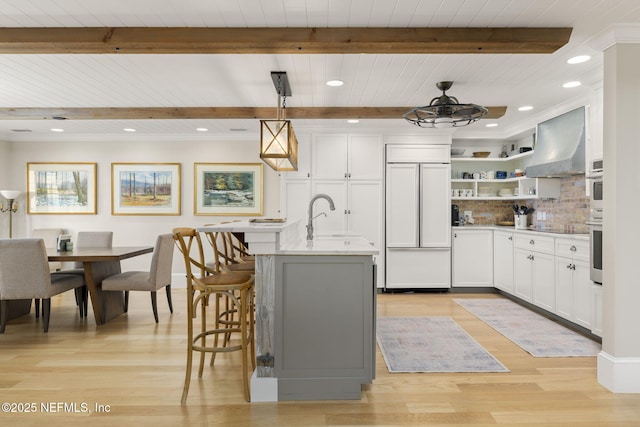 kitchen with white cabinetry, pendant lighting, an island with sink, paneled fridge, and wall chimney exhaust hood