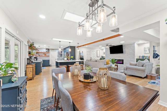 dining space with a skylight, ceiling fan, crown molding, light hardwood / wood-style flooring, and a stone fireplace