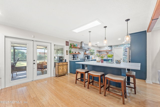 kitchen with french doors, wall chimney exhaust hood, light hardwood / wood-style floors, hanging light fixtures, and a breakfast bar area