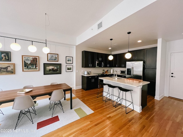 kitchen featuring stainless steel appliances, an island with sink, light hardwood / wood-style floors, decorative light fixtures, and a breakfast bar area