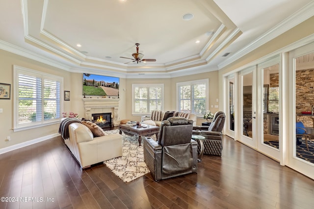 living area featuring a tray ceiling, dark wood-style flooring, crown molding, a glass covered fireplace, and ceiling fan