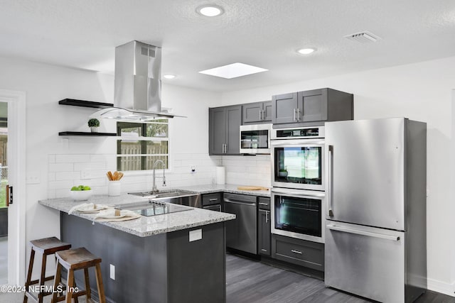 kitchen featuring a skylight, light stone countertops, appliances with stainless steel finishes, kitchen peninsula, and island exhaust hood