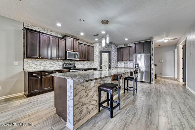 kitchen with tasteful backsplash, light stone counters, decorative light fixtures, a center island with sink, and appliances with stainless steel finishes