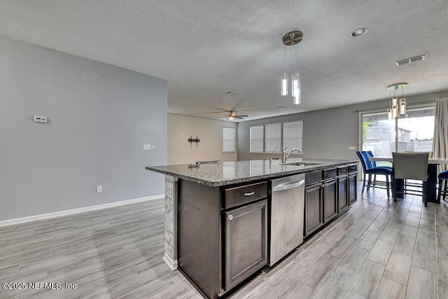 kitchen featuring light stone countertops, stainless steel dishwasher, dark brown cabinets, pendant lighting, and an island with sink