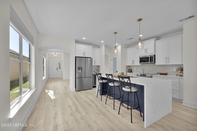 kitchen with decorative backsplash, appliances with stainless steel finishes, decorative light fixtures, white cabinetry, and an island with sink