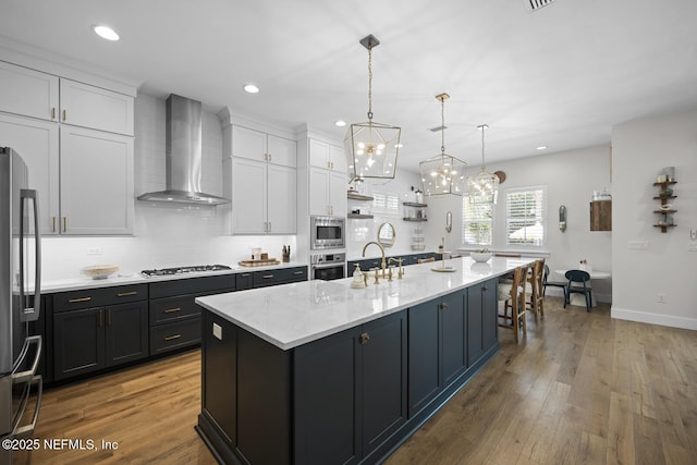 kitchen with white cabinets, a center island with sink, wall chimney range hood, light wood-type flooring, and stainless steel appliances