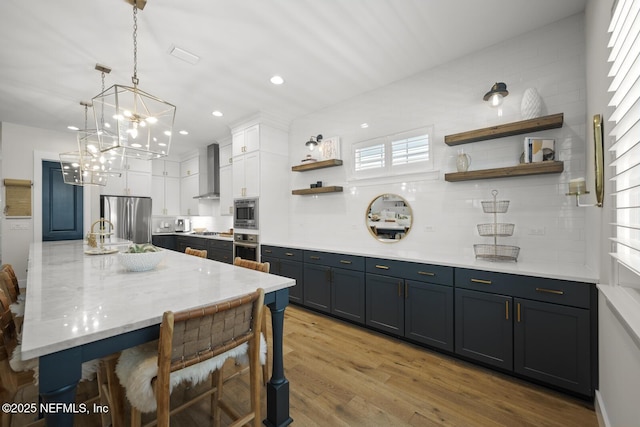 kitchen featuring stainless steel appliances, wall chimney range hood, light hardwood / wood-style flooring, pendant lighting, and white cabinets