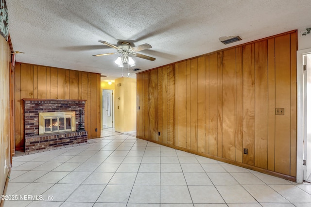 unfurnished living room featuring ceiling fan, a brick fireplace, a textured ceiling, wooden walls, and light tile patterned flooring