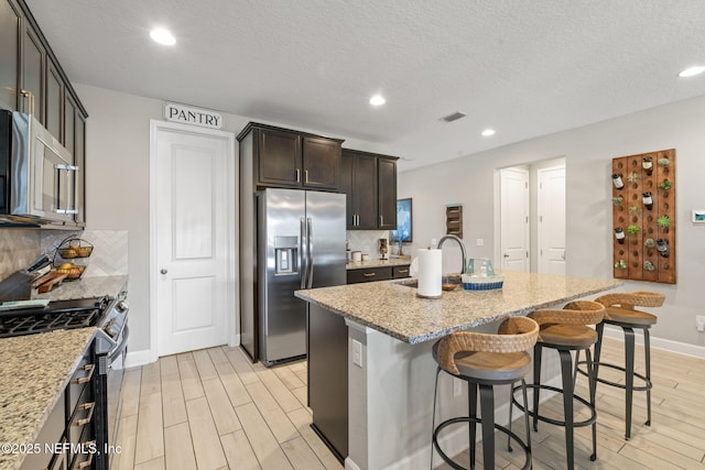 kitchen with appliances with stainless steel finishes, backsplash, light stone counters, and a kitchen island with sink