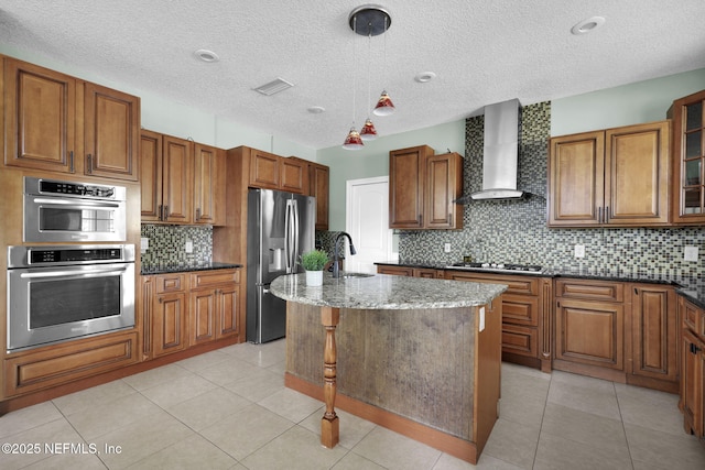 kitchen featuring backsplash, dark stone counters, a center island with sink, wall chimney range hood, and stainless steel appliances