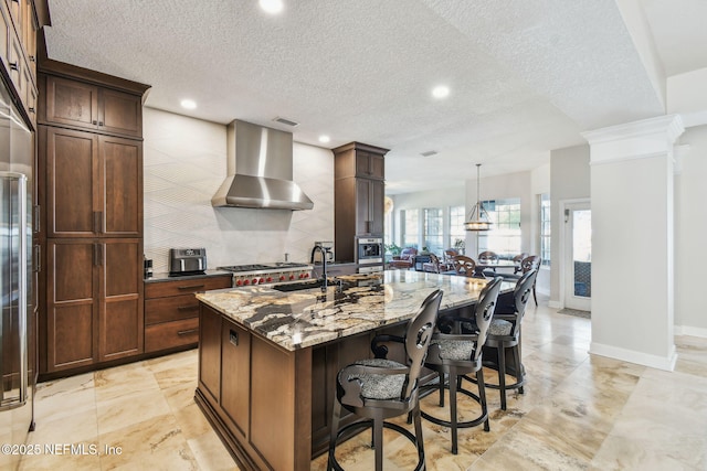 kitchen with sink, wall chimney range hood, light stone counters, decorative light fixtures, and a center island with sink