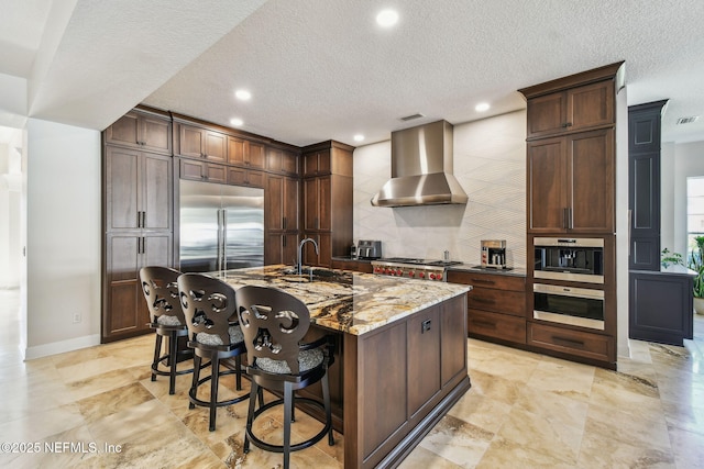 kitchen with sink, wall chimney exhaust hood, light stone countertops, an island with sink, and stainless steel appliances