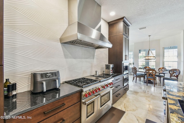 kitchen featuring wall chimney exhaust hood, hanging light fixtures, dark stone counters, a textured ceiling, and appliances with stainless steel finishes