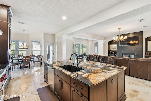 kitchen with dark stone counters, a center island with sink, sink, stainless steel dishwasher, and a large fireplace